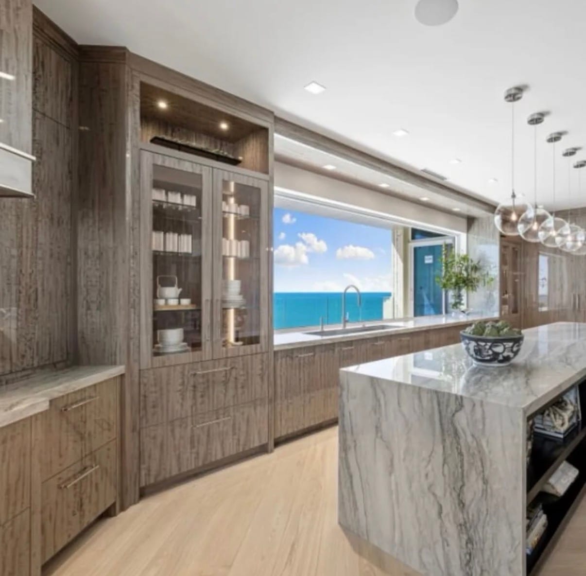 A coastal kitchen in grey-dyed figured eucalyptus veneer with a marble island, the sea visible through the window behind